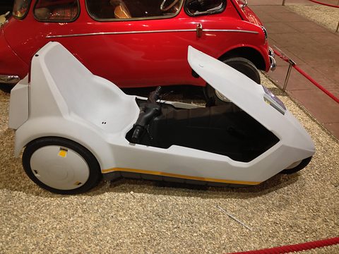 White electric tricycle side-on on gravel, a red bubble car behind it. Opens a larger view