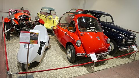 Red bubble car on a gravel display behind a rope, with a white Sinclair C5, a yellow bubble car and a dark saloon. Opens a larger view