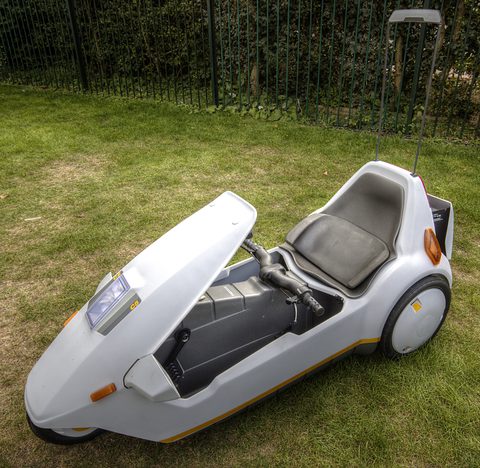 White electric tricycle on grass with a tall thin mast rising from behind the seat.