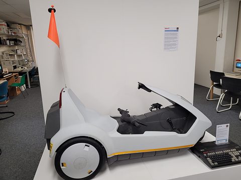 White electric tricycle with a tall orange flag, on a plinth beside a black computer keyboard. Opens a larger view