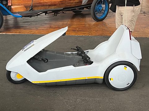 White electric tricycle side-on on a dark floor, a blue-wheeled veteran car behind. Opens a larger view