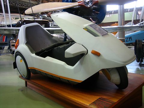 White electric tricycle on a wooden plinth, the wing of a biplane overhead. Opens a larger view