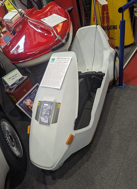 White electric tricycle seen from the front quarter in a museum, a placard on its upturned nose. Opens a larger view
