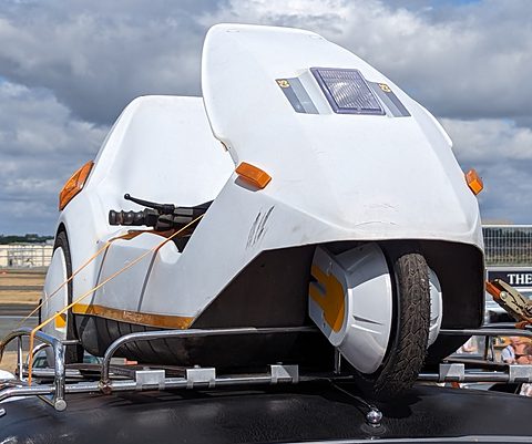 White electric tricycle tied with orange straps onto a chrome roof rack, under a cloudy sky. Opens a larger view