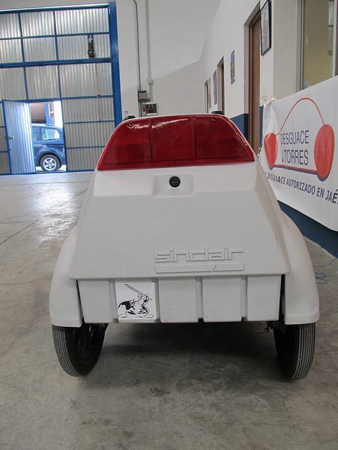 White electric tricycle from the rear, a red tail lamp above the moulded Sinclair lettering, in a hall. Opens a larger view