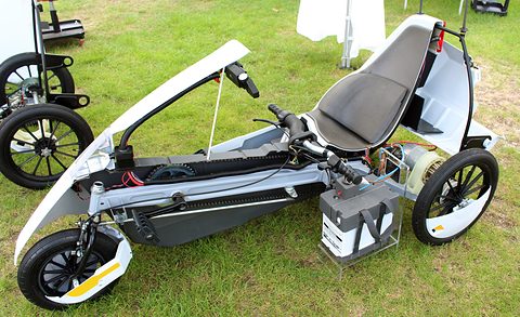 Cut-away electric tricycle on grass with part of its white body removed to show the frame and battery.