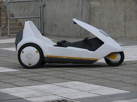 White electric tricycle side-on on a chequered pavement in front of a concrete wall. Opens a larger view