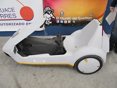 White electric tricycle side-on in front of a banner with pixel-art characters. Opens a larger view