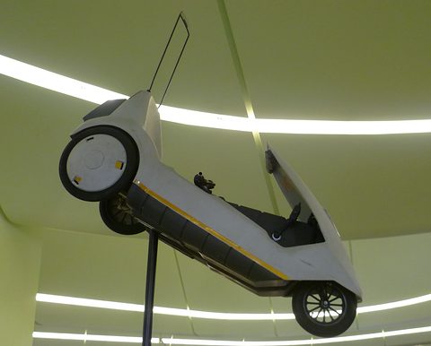 White electric tricycle tilted on a pole against a lime-lit museum ceiling.