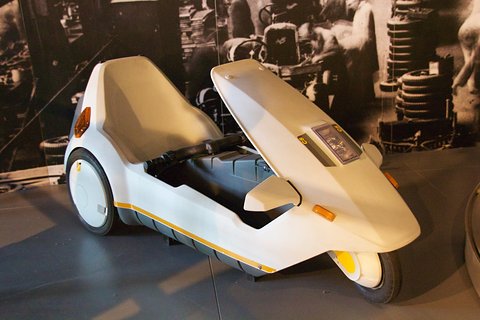 White electric tricycle in front of a large black-and-white factory photograph. Opens a larger view