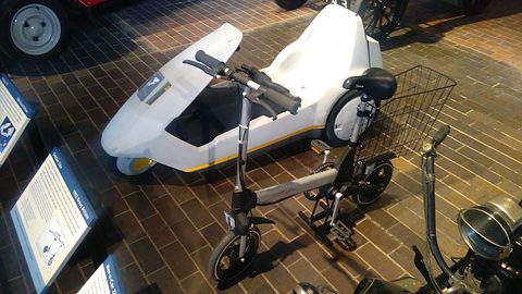 Small folding electric bicycle with a basket in front of a white electric tricycle on a brick floor. Opens a larger view