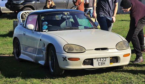 White small two-seater with oval headlamps and a K422 plate on grass, people standing beside it. Opens a larger view