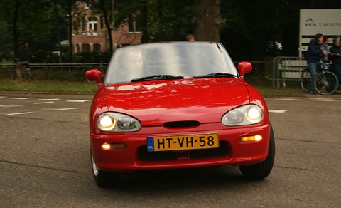 Front of a red small two-seater with its headlamps lit and a yellow Dutch plate on a road. Opens a larger view