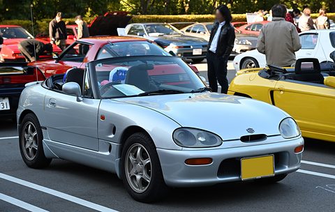 Silver small open two-seater with oval headlamps and a yellow plate in a car park, people behind. Opens a larger view