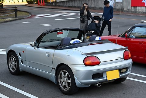 Rear of a silver small open two-seater with a boot spoiler and yellow plate, two people with a pushchair behind. Opens a larger view