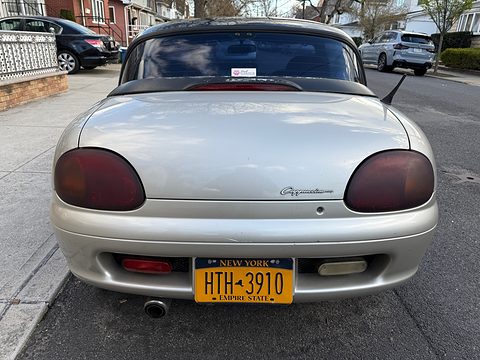 Rear of a silver small roadster with large round tail lamps and a yellow New York plate in a street. Opens a larger view