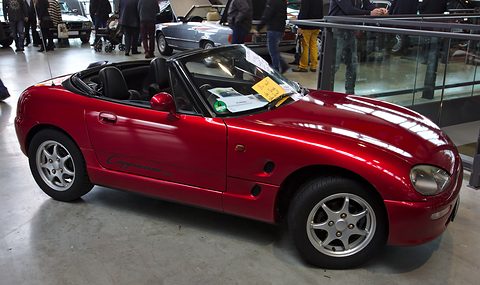 Red small open two-seater with a for-sale notice behind the windscreen on a show-hall floor.