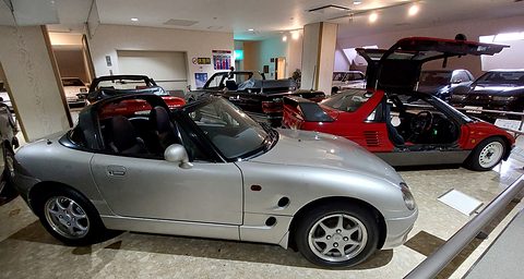 Side view of a silver small open two-seater in a museum hall, a red mid-engined coupé with doors raised behind. Opens a larger view