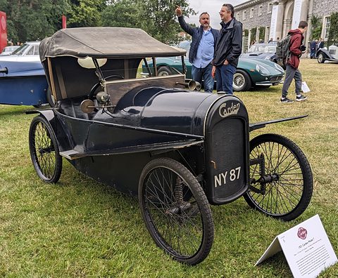 A small black open cyclecar with a raised fabric hood, wire wheels and Carden script on its nose, spectators behind. Opens a larger view