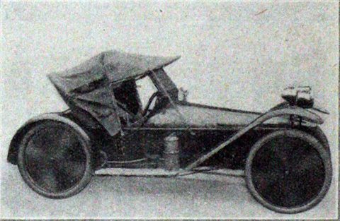 Side view of a small single-seat cyclecar with a fabric hood, disc wheels and an engine and fuel tank over the tail.