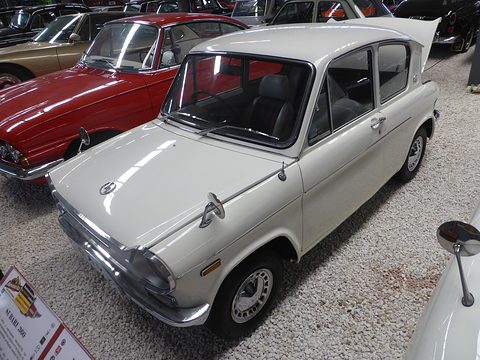 White two-door small saloon on a gravel floor beside a red car, its rear window seen undercut. Opens a larger view