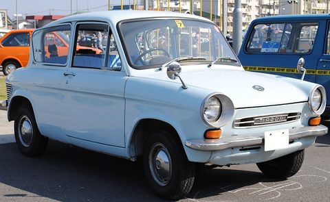 Pale blue small two-door saloon with round headlamps and a chrome nose, parked beside a blue van. Opens a larger view
