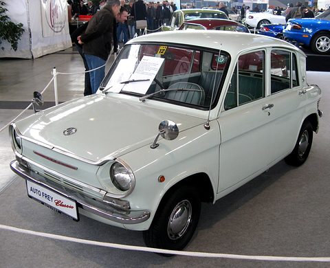 White four-door saloon with a display card on its windscreen, a blue sports car behind. Opens a larger view