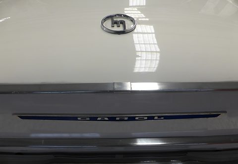 Close view of a white bonnet with a chrome Mazda m badge above a strip lettered CAROL. Opens a larger view