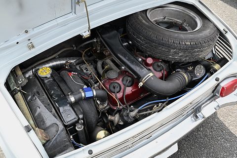 Open rear lid of a white car showing a compact engine, hoses and a spare tyre on top.