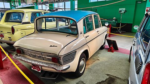 Rear three-quarter of a cream saloon with a blue roof and louvred tail, green museum walls behind. Opens a larger view