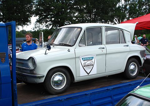 White four-door saloon with a shield emblem on the door, standing on a blue trailer under trees. Opens a larger view