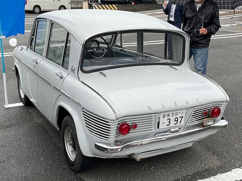 Rear three-quarter of a pale grey four-door saloon with a louvred tail and reverse-rake window. Opens a larger view