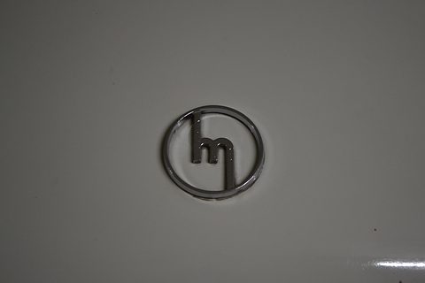 A round chrome badge with a stylised lower-case m on a plain grey bonnet. Opens a larger view
