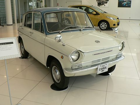 Cream four-door small saloon on a polished white floor, a yellow modern Mazda behind it. Opens a larger view