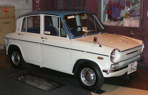 White four-door saloon with a blue roof on a museum floor, a red-brown wall behind. Opens a larger view