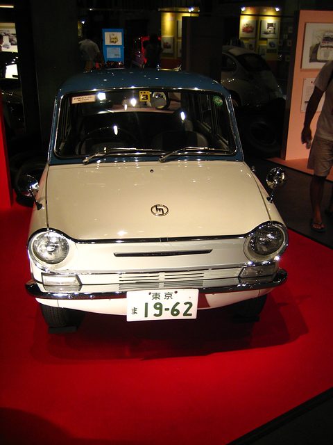 Front of a white saloon on a red stand, round headlamps and a Tokyo number plate, in an upright frame. Opens a larger view