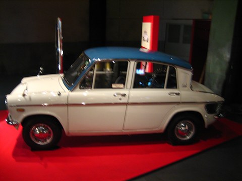 Side view of a white saloon with a blue roof on a red carpeted stand against a dark background. Opens a larger view