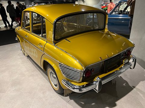 Rear three-quarter of a gold saloon with a chrome louvred tail, visitors in the background. Opens a larger view