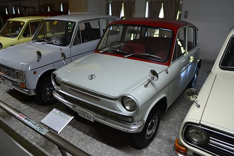 White four-door saloon with a red roof in a museum row behind a low rail, grey cars on either side. Opens a larger view