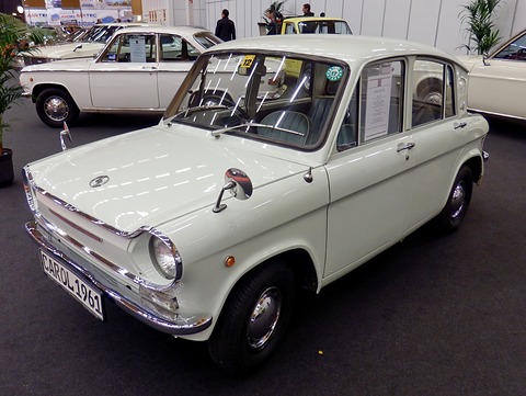 White four-door small saloon on a dark carpet, with a German-style number plate and cars behind. Opens a larger view