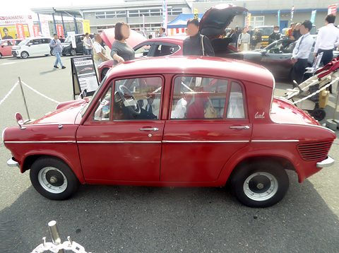 Side view of a red four-door saloon showing the reverse-rake rear window, visitors behind. Opens a larger view