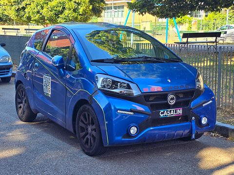 Blue modern two-door microcar with slim headlamps and a white CASALINI plate on its nose, parked under trees.