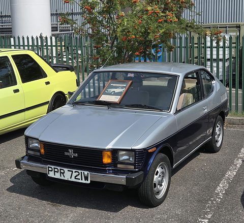 Silver and dark blue small fastback with square headlamps and a British plate, on a car park beside a yellow hatchback.