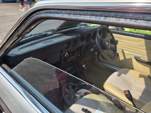 Small car interior through an open window: dark dashboard with round dials, a steering wheel and beige seats. Opens a larger view