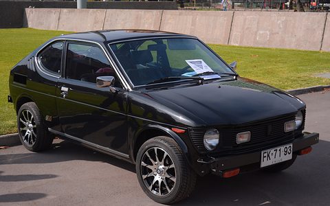 Black small fastback coupé with round headlamps, aftermarket multi-spoke wheels and a Chilean plate, parked by a lawn. Opens a larger view