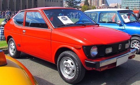 Red small fastback coupé with round headlamps and a black grille, a notice on its dashboard, a blue car and tower blocks behind. Opens a larger view