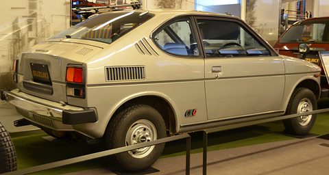 Rear quarter of a silver small fastback with louvres behind the side window and a CX-G badge, behind a rail on a museum floor. Opens a larger view
