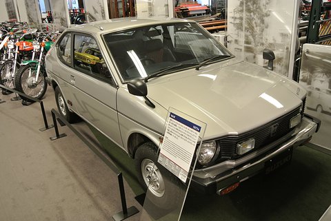 Silver small fastback coupé with round headlamps and a black grille behind an information placard, motorcycles on display alongside.