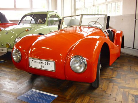 Orange open two-seater with a “Champion 250 1950” plate on a parquet floor, a green Champion behind. Opens a larger view