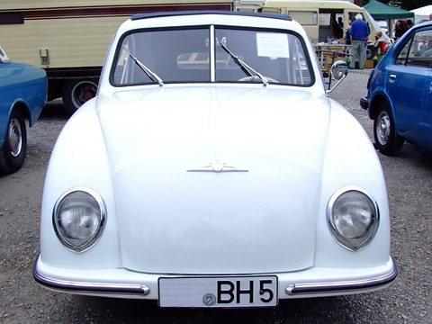 White small car facing the camera with a winged nose badge and round headlamps on gravel, stalls behind. Opens a larger view